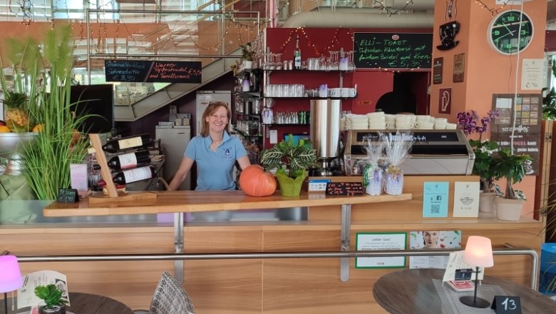Cafe Elli, © Cafe Elli Eine Frau steht hinter der Theke eines Cafés mit Pflanzen, Weinflaschen und einer Kaffeemaschine.