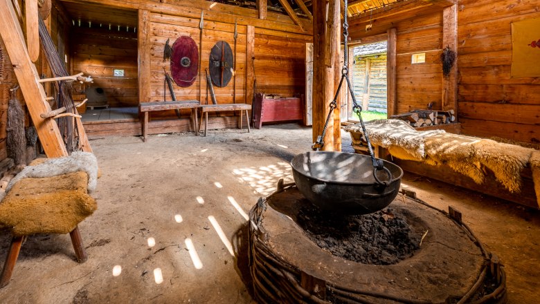 Celtic village Schwarzenbach, © Wiener Alpen, Christian Kremsl Interior view of a reconstructed Celtic house with wooden walls, a large cauldron over a fireplace and shields on the wall.