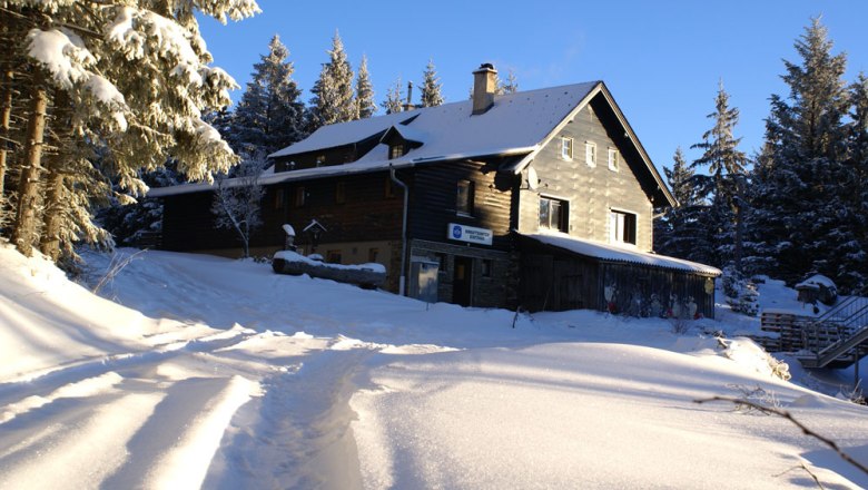 Winterimpression - Herrgottschnitzerhütte, © Herrgottschnitzer-Franz-Kaupe-Haus, Mimm Verschneite Hütte im Wald bei Sonnenschein.
