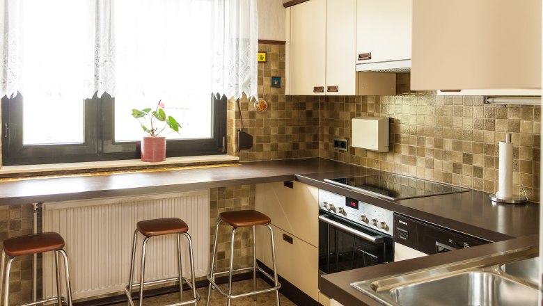 Küche, © Fam. Sulzbacher Moderne Küche mit braunen Fliesen, Herd, Spüle und Barhockern vor einem Fenster mit Vorhängen.