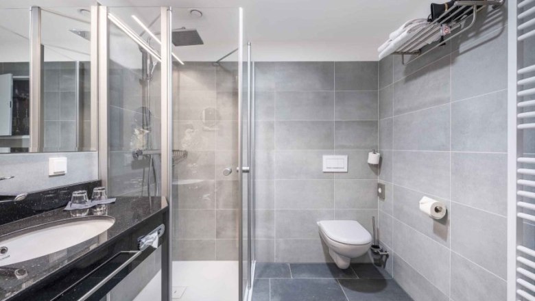 PLAZA Residence Wiener Neustadt Badezimmer, © PLAZA Residence Wiener Neustadt Modernes Badezimmer mit grauen Fliesen, Dusche, Waschbecken und WC.