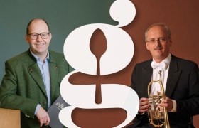 s'Hutwisch mit Solotrompeter a. D. der Wiener Philharmoniker Hans-Peter Schuh, © Wiener Alpen/Sophie Menegaldo, Hans-Peter Schuh Zwei Männer in Anzügen mit einem großen stilisierten Buchstaben im Vordergrund.