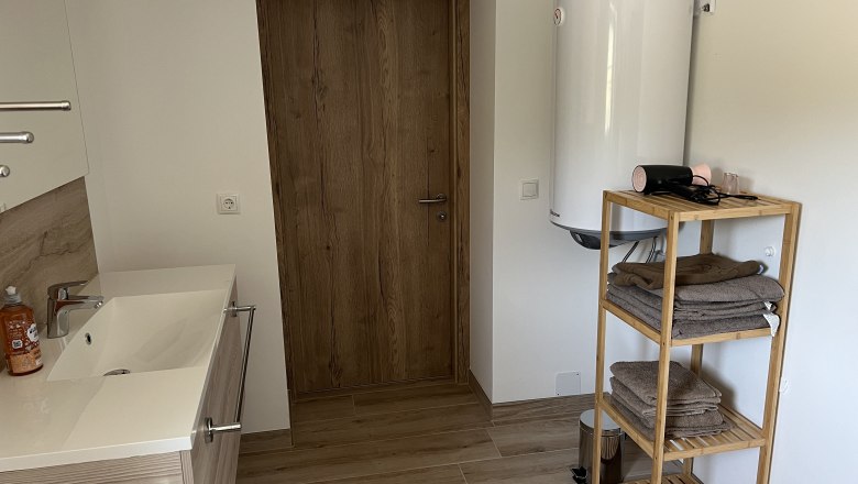 Badzimmer, © Anton Sommer Modernes Badezimmer mit Holzregal, Handtüchern, Waschbecken und Warmwasserboiler.