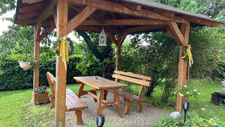 Laube im Garten, © Wiener Alpen Holzlaube im Garten mit Tisch und Bänken, umgeben von Pflanzen.