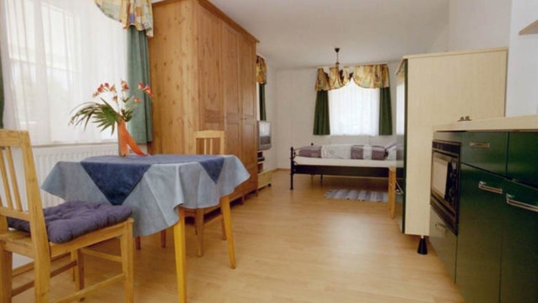 Wohnung 30m², © Haus Lenz Kleine Wohnung mit Holzmöbeln, Esstisch, Stühlen, Bett und Küchenzeile.