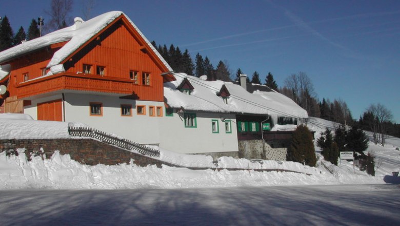 Gasthaus Dissauer am Feistritzsattel, © Tourismusbüro Kirchberg/Wechsel Ein verschneites Gasthaus mit rotem Holzgiebel und weißen Wänden am Feistritzsattel, umgeben von Bäumen und blauem Himmel.