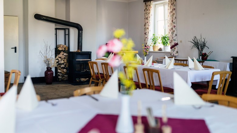 Saal im Gasthaus der Kräuterwirtin, © Nicole Heiling Gemütlicher Gastraum mit Holzofen, gedeckten Tischen und Blumen.