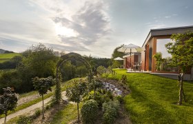 Wiener Alpen Bett: "Bett in da Wies´n", © Bene Croy Moderne Unterkunft in grüner Landschaft mit Sonnenschirmen und Garten.