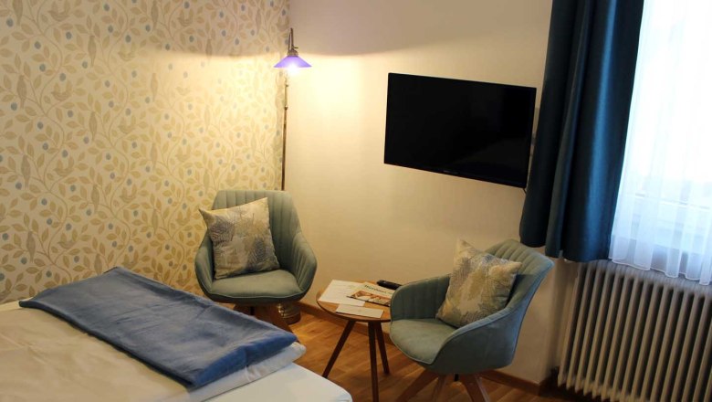 Zimmer mit Bett, Tisch mit zwei Stühlen, Fernseher, Fenster mit blauen Vorhängen und Stehlampe., © Magdalena Donhauser Zimmer mit Bett, Tisch mit zwei Stühlen, Fernseher, Fenster mit blauen Vorhängen und Stehlampe.