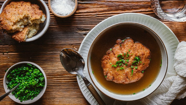 Cheese dumpling soup, © Koll GmbH Cheese dumpling soup in a bowl on a wooden table, with chives and salt in small bowls next to it.
