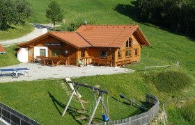 Simashütte, © Birgit Gruber Eine Berghütte namens Simashütte mit rotem Dach, umgeben von grünen Wiesen und einem Spielplatz mit Schaukel.