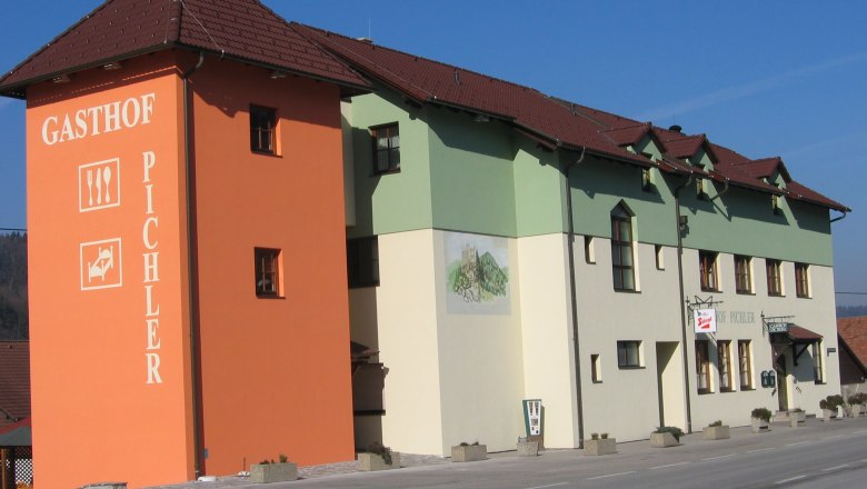 Haus, © Peter Pichler Ein Gasthof mit orangefarbenem Turm und grün-weißer Fassade an einer Straße.
