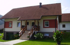 Ferienwohnung Maria Grabner, © Stadtgemeinde Gloggnitz Ein Einfamilienhaus mit rotem Dach und weiß-roter Fassade, umgeben von gepflegtem Garten und Blumen.