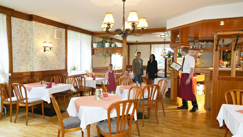 Rustikaler Charme in der Stube, © Hausmann/Zwickl Ein rustikales Restaurant mit Holzmöbeln und Tischdecken. Ein Kellner begrüßt eine Familie am Eingang.