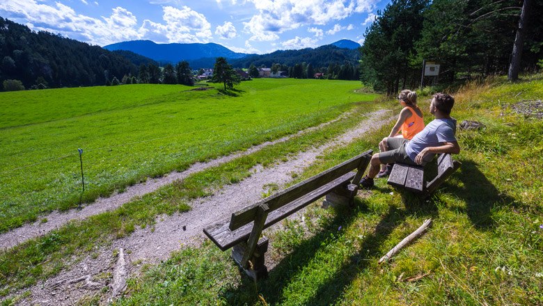 Ausblick in die Gegend rund um Rohr, © Wiener Alpen, Christian Kremsl Zwei Personen sitzen auf einer Bank und blicken auf eine grüne Wiese mit Bergen im Hintergrund.