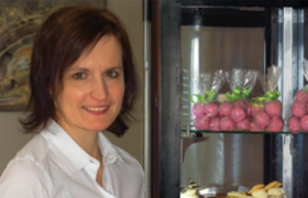 Kaffeehaus Siaß´ Eck, © Sooo gut schmeckt die Bucklige Welt Eine Frau mit weißer Bluse steht lächend neben einer Vitrine mit verpackter Mehlspeise.