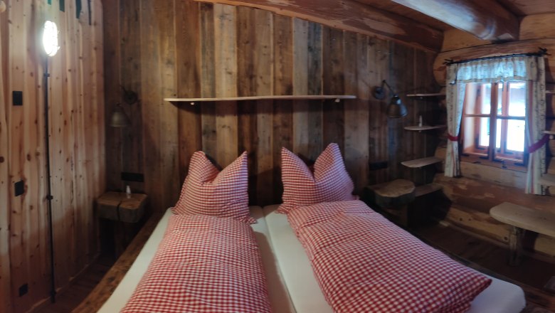 Zimmer, © 2021 Maxsam GmbH Rustikales Zimmer mit Holzwänden, Bett mit rot-weiß karierten Kissen, Fenster mit Vorhängen, Holztür und Regalen.