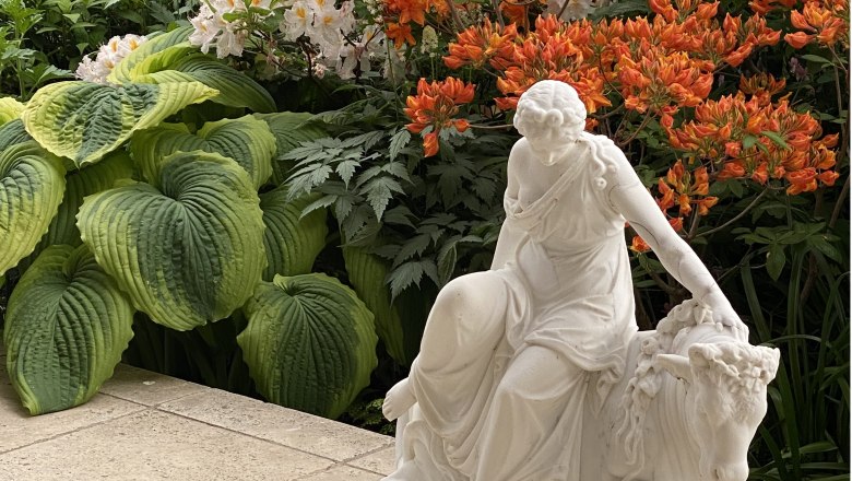Fernery - Glashaus im Ecolution.Lab, © Bernd Hochwartner Weiße Statue einer Frau neben großen grünen Blättern und orangefarbenen Blumen.