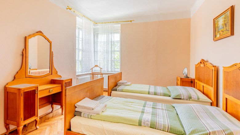 Bedroom, © Wiener Alpen / Christian Kremsl A traditional bedroom with two single beds, a dressing table with mirror and a window with white curtains.
