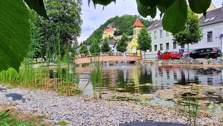 Blick auf die Bergkirche Pitten, © Wiener Alpen Blick auf einen Teich mit Brücke und Bergkirche im Hintergrund, umgeben von Bäumen und Häusern.