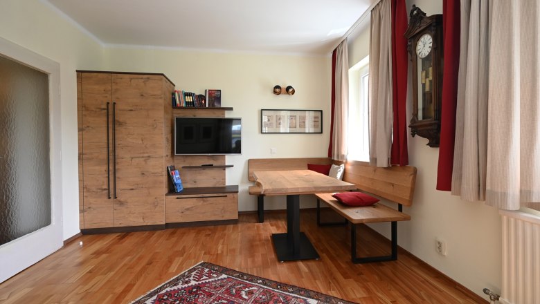 Wohnzimmer, © Christine Kuppelwieser Gemütliches Wohnzimmer mit Holzmöbeln, einem Teppich, einer Eckbank und einer Wanduhr.