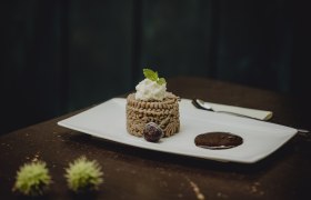 Ganzjähriger Favorit: Kastanienmousse, © Niederösterreich-Werbung/Sophie Menegaldo Ein Dessert aus Kastanienmousse auf einem weißen Teller, garniert mit Sahne und Minzblatt, daneben eine Schokoladensauce und eine Kirsche.