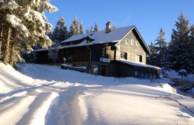 Winterimpression - Herrgottschnitzerhütte, © Herrgottschnitzer-Franz-Kaupe-Haus, Mimm Verschneite Hütte im Wald bei Sonnenschein.