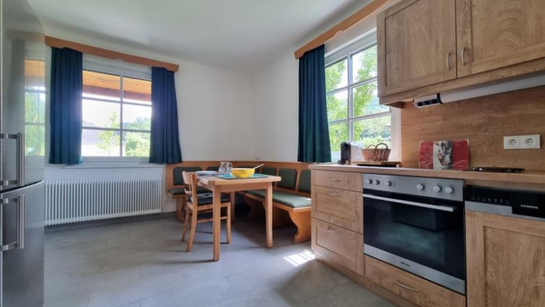 Apartment G6 im Gerhardthof Reichenau, © Gerhardthof Reichenau Küche mit Holzmöbeln, Esstisch und Eckbank, zwei Fenstern mit grünen Vorhängen.