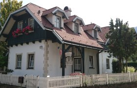 Landhaus Scheibenreif, © Scheibenreif Karin Ein traditionelles Landhaus mit Holzverkleidung, roten Dachziegeln und Blumenkästen vor den Fenstern. Ein weißer Holzzaun umgibt das Grundstück.