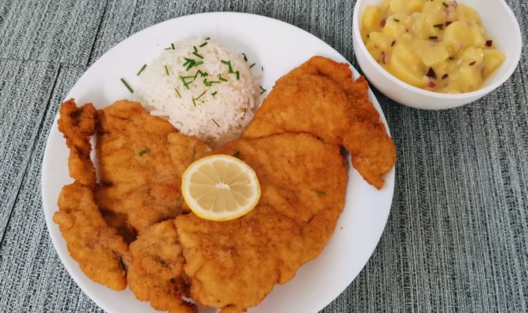 Cafe Elli, © Cafe Elli Ein Teller mit paniertem Schnitzel, Reis und einer Zitronenscheibe, daneben eine Schüssel Kartoffelsalat.