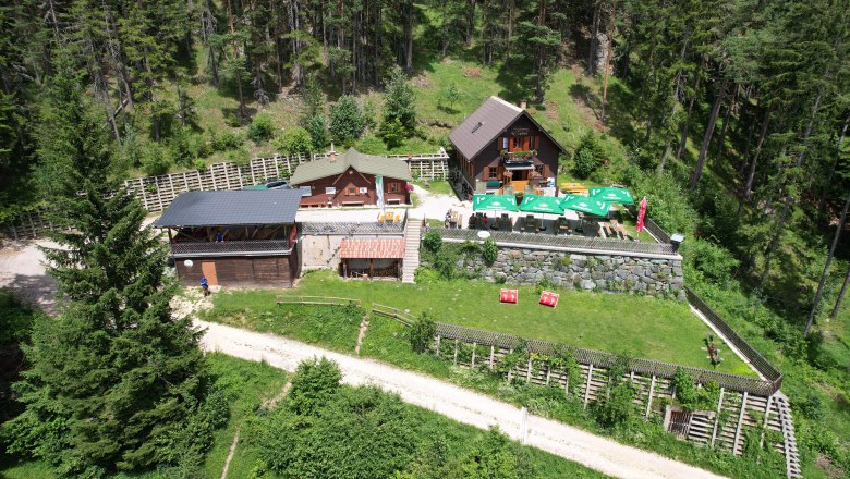 Pottschacherhütte, © Georg Eder Luftaufnahme der Potschacherhütte in einem Waldgebiet mit umliegenden Bäumen und einer Terrasse mit Sonnenschirmen.
