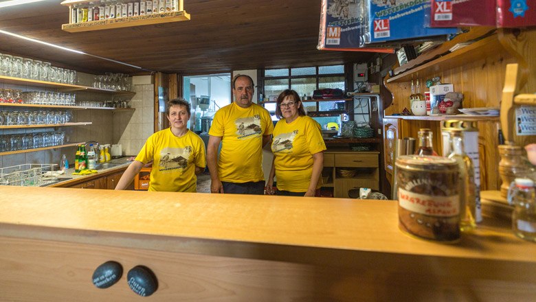 The team at the Wetterkoglerhaus, © Wiener Alpen, Christian Kremsl Three people in yellow T-shirts are standing behind a bar in a rustic room.