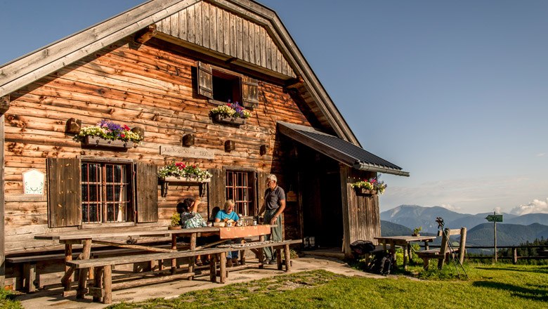 The Kranichberger Schwaig, © Niederösterreich-Werbung, Robert Herbst Wooden hut in the mountains with people at the table, blue sky.