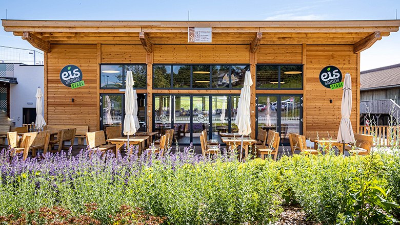 Eis-Greissler Kulinarik-Stadl, © Blochberger Eisproduktion GmbH Modernes Holzgebäude mit Terasse, auf der Bänke, Stühle und Sonnenschirme stehen.
