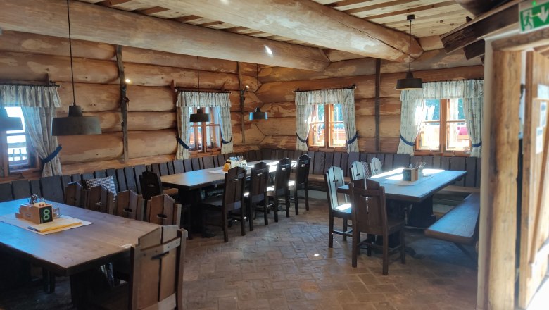 Gemütliches Ambiente in der Stoa Alm, © 2021 Maxsam GmbH Innenansicht einer rustikalen Holzhütte mit Holztischen und Stühlen, dekoriert mit Vorhängen an den Fenstern.
