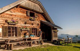 Die Kranichberger Schwaig, © Niederösterreich-Werbung, Robert Herbst Holzhütte in den Bergen mit Menschen am Tisch, blauer Himmel.