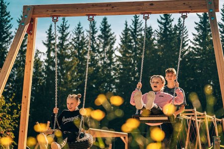 Riesenschaukel am Hirschi-Spielplatz, © Bergbahnen Semmering Hirschenkogel GmbH Drei Kinder schaukeln auf einer großen Schaukel im Freien, umgeben von Bäumen.
