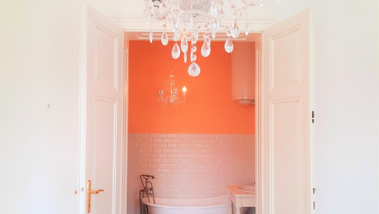 Einblick in das Badezimmer, © Sisi-Schloss Rudolfsvilla Blick in ein Badezimmer mit freistehender Badewanne, orangefarbener Wand und Kronleuchter.
