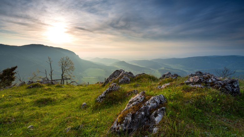 Ausblick von der Geländehütte, © Wiener Alpen, Christian Kremsl Panoramablick von einem Hügel mit Felsen und Gras im Vordergrund, Bergen und einem bewölkten Himmel im Hintergrund.
