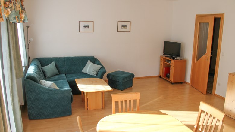 Wohnung 70m², © Haus Lenz Wohnzimmer mit grüner Eckcouch, Holztisch, Fernseher und Esstisch.