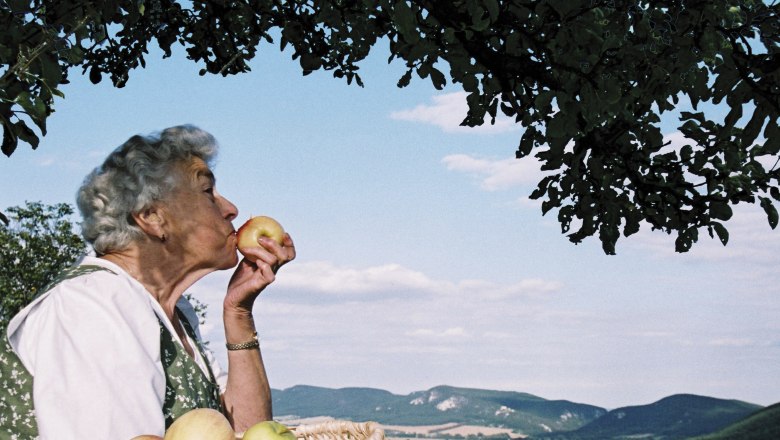 Mohr-Sederl Fruit World, © Mohr-Sederl Fruchtwelt Elderly woman with a basket full of apples kissing an apple in front of a landscape with hills and blue sky.