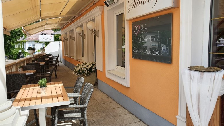 Herzlich Willkommen im Stadtcafé, © Stadtcafé Gloggnitz Außenbereich eines Cafés mit Tischen und Stühlen, orangefarbene Wand mit Schild 'Stadtcafé'.