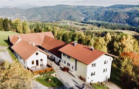 Koglhof St. Corona, © Andrea Tauchner Luftaufnahme eines Bauernhofs mit roten Dächern in einer hügeligen Landschaft.