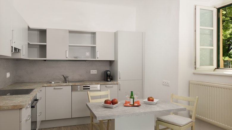 Küche, © Landhaus Puchbergerhof by smc Moderne Küche mit grauen Schränken, Tisch mit Äpfeln und Getränk, offenes Fenster.