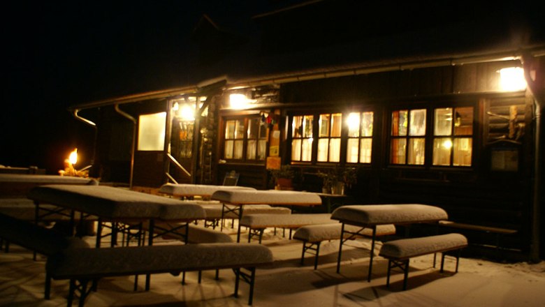 winternacht-herrgottschnitzerhuette, © Herrgottschnitzer-Franz-Kaupe-Haus, Mimm Verschneite Bänke vor einer beleuchteten Hütte in der Nacht.