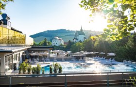 Vivea 4* Hotel Bad Schönau Zum Landsknecht, © Hannes Dabernig Außengelände eines Hotels mit Pool, Sonnenschirmen und Liegen, umgeben von Natur und Kirche im Hintergrund.