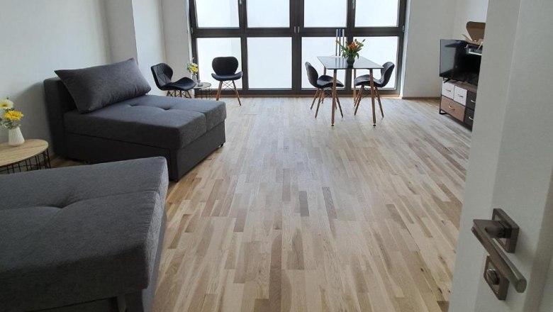 Green Point Apartments, © Green Point Apartments Modernes Zimmer mit Holzboden, grauen Sofas, Esstisch und großen Fenstern.