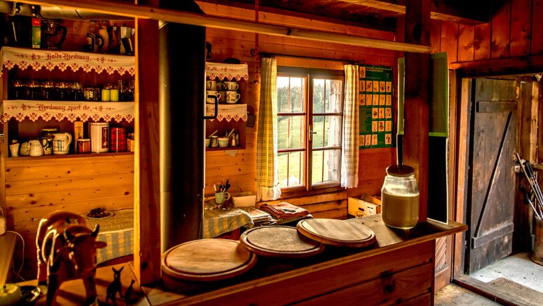 The cozy parlor of the Kranichberger Schwaig, © Niederösterreich-Werbung, Robert Herbst Cozy wooden hut with rustic furnishings, a window with curtains and an open door area.