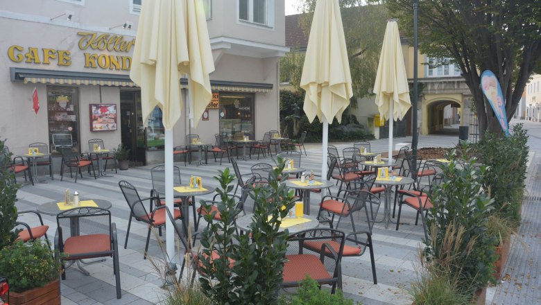 Pub garden, © Konditorei Köller An empty pavement garden with tables and chairs in front of a café with yellow parasols.