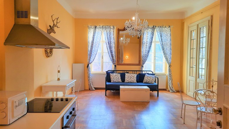 Salon mit Küche und Essplatz, © Sisi-Schloss Rudolfsvilla Wohnzimmer mit Küche, Esstisch und Sofa, gelbe Wände, Kronleuchter, Fenster mit Vorhängen.